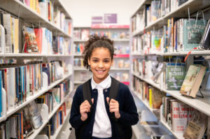 Female child with backpack standing between bookshelves